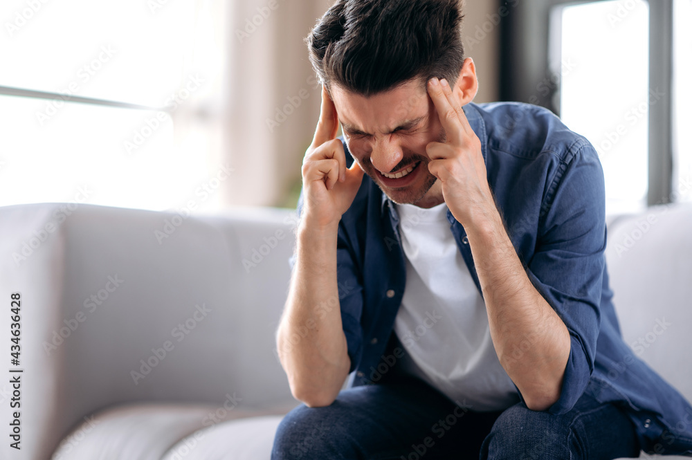 Headache, migraine. Caucasian young adult guy sitting at home on the sofa in the living room, massaging temples, suffering from headache, migraine, stress Headache, migraine. Caucasian young adult guy sitting at home on the sofa in the living room, massaging temples, suffering from headache, migraine, stress