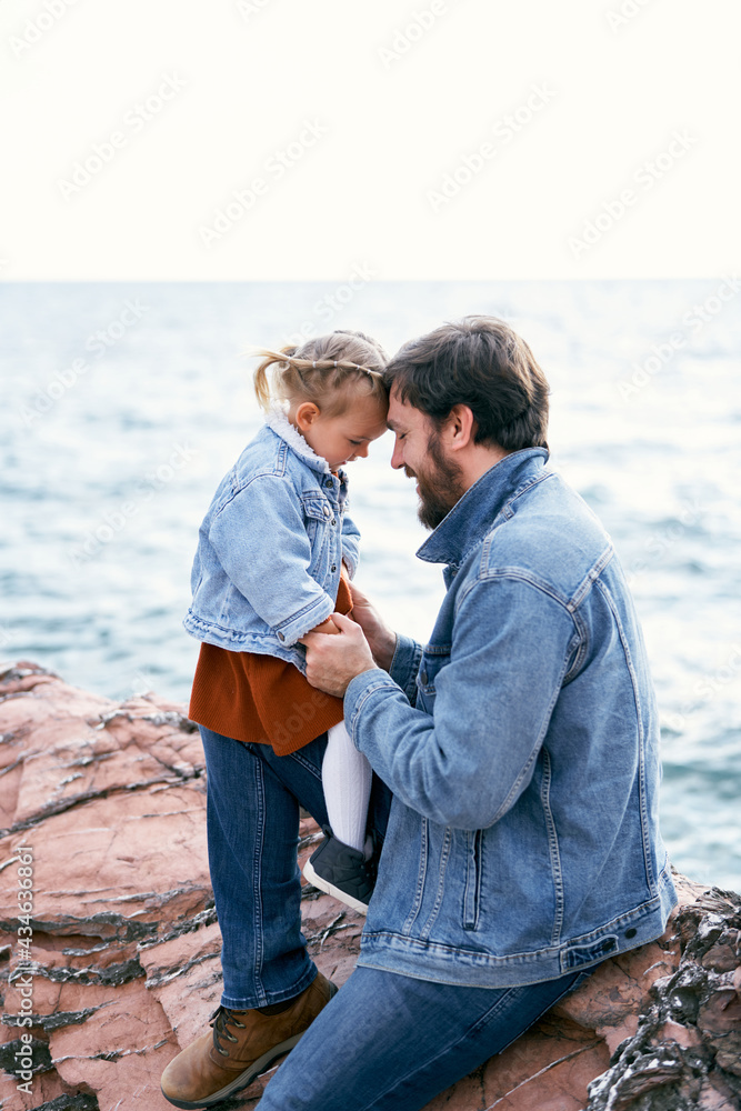 Little girl sits on daddy's feet, bowing her forehead to his forehead. Daddy sits on stones against the background of the sea