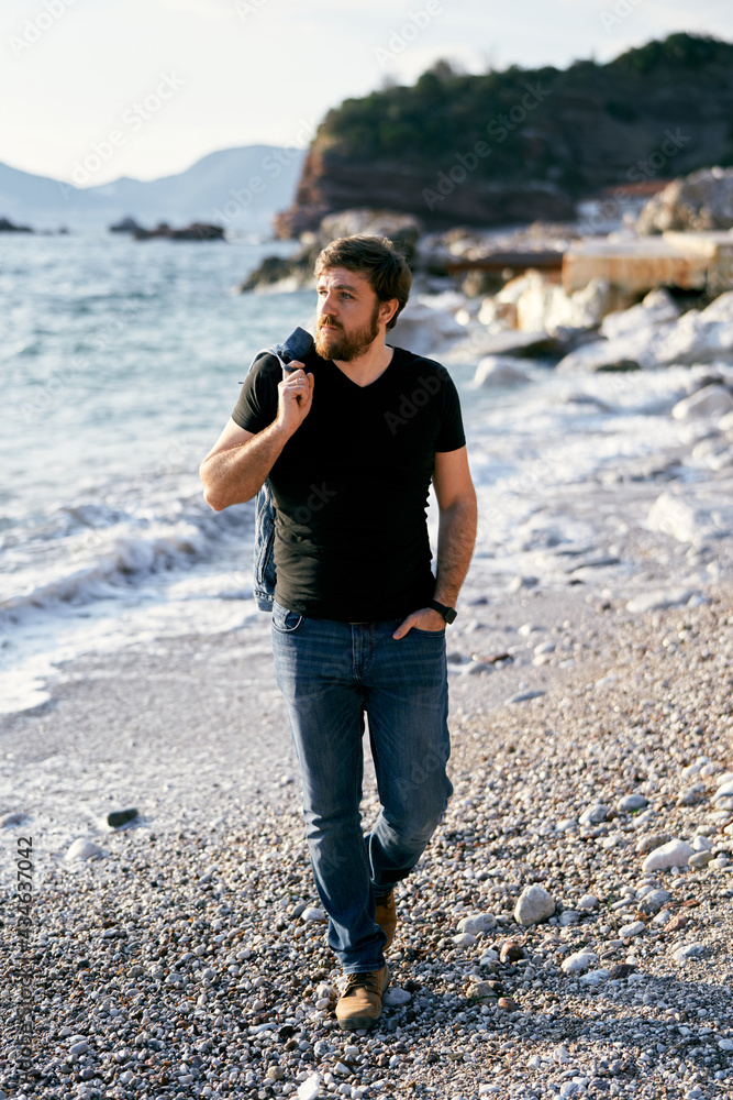 Pensive man walking along a pebble beach with a denim jacket over his shoulder and a hand in his pocket against the background of the sea and rocks