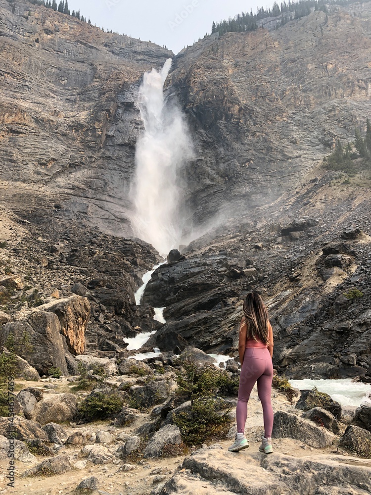 Obraz premium A pretty female hiker in the pink athletic outfit is posing in front of Takakkaw Falls in Yoho National Park, British Columbia, Canada