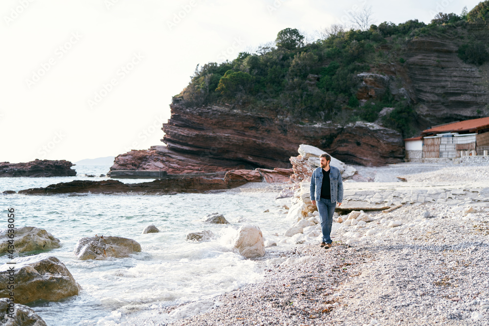 Pensive man in jeans and a denim jacket walks along a pebble beach looking at the water against a background of green rocks