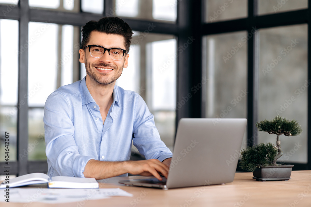 Portrait of a handsome intelligent caucasian man with glasses, businessman or manager, in formal shirt, sitting at a table with laptop, looking at camera and smiling friendly Portrait of a handsome intelligent caucasian man with glasses, businessman or manager, in formal shirt, sitting at a table with laptop, looking at camera and smiling friendly