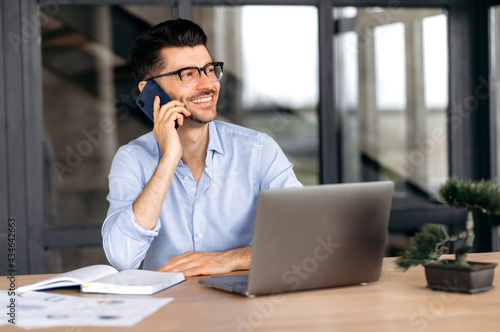 Mobile communication concept. Smiling caucasian freelancer talking on cellphone while working on laptop, positive man in formal shirt having pleasant phone conversation, smiling friendly