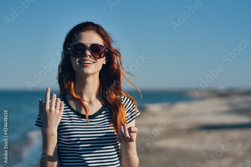 woman in sunglasses and a striped t-shirt gestures with her hands in the mountains near the sea on the beach