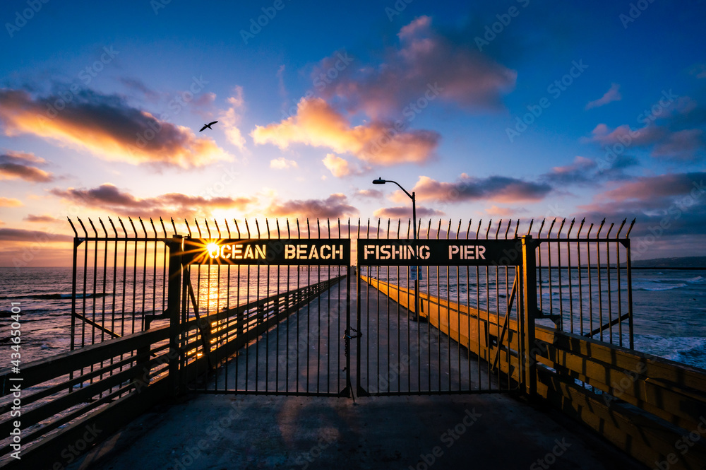 Ocean beach fishing pier in San Diego California during colorful sunset with a sun star and bird flying in the sky