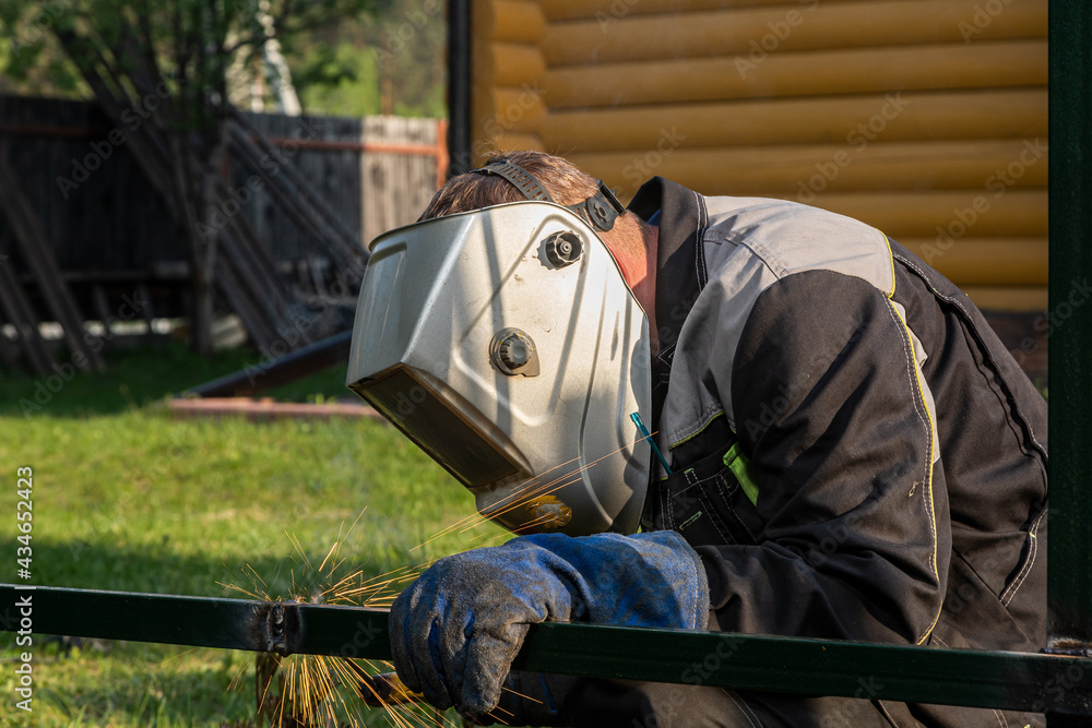 Close up of a young  man welder in  uniform, welding mask and welders leathers, weld  metal  with a  welding machine at the construction  of a fence in summer day putside in the village