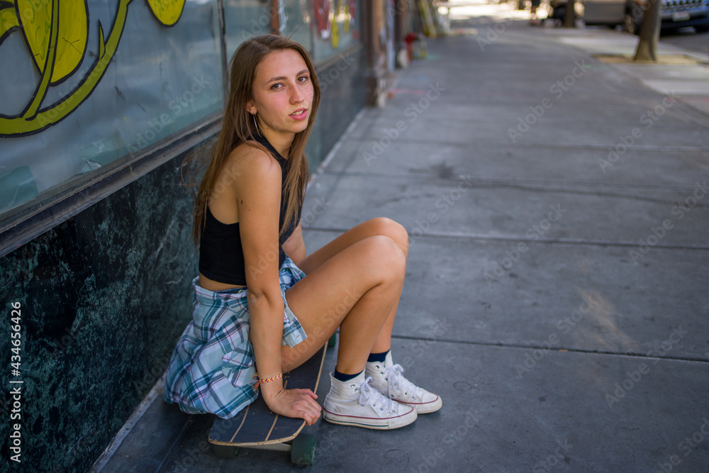 Naklejka premium Young Woman Seated With Skateboard on the Sidewalk in a Downtown Area