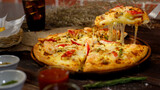 Selective focus and close up on a piece of double cheese homemade seafood pizza on spatula or scoop with blur background of wooden table, cold beverage and brick wall. Food Concept.