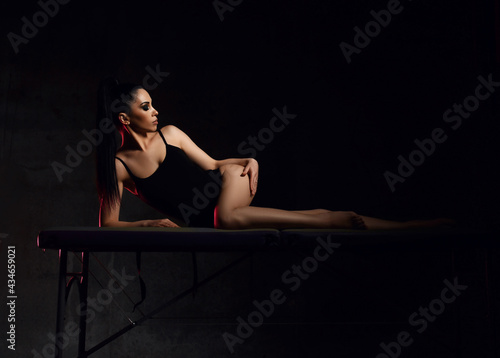 Tempting alluring young sexy brunette woman in black bodysuit is lying on massage table, touching her smooth silky skin on hip after cosmetic procedure, anti-cellulite therapy over dark background