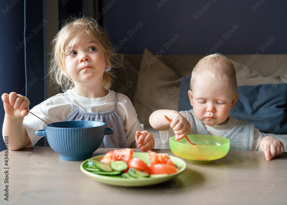 Two small cute kids older sister and baby boy brother having healthy lunch, eating soup and fresh vegetables cucumbers tomatoes while sitting at wooden table at dark home and enjoying vegetarian meal Two small cute kids older sister and baby boy brother having healthy lunch, eating soup and fresh vegetables cucumbers tomatoes while sitting at wooden table at dark home and enjoying vegetarian meal