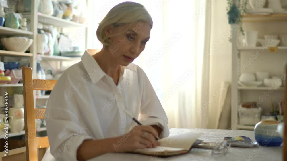 Aged woman writing information in textbook planner spending daytime in own art studio, serious female with notepad checking time on wearable smartwatch and using cellular device for web messaging Aged woman writing information in textbook planner spending daytime in own art studio, serious female with notepad checking time on wearable smartwatch and using cellular device for web messaging