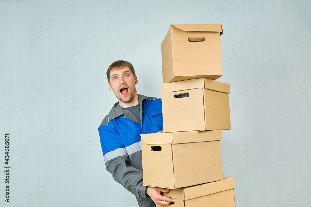 A happy man in overalls looks out from behind a pile of cardboard boxes. A professional mover performs the transfer. A happy man in overalls looks out from behind a pile of cardboard boxes. A professional mover performs the transfer.