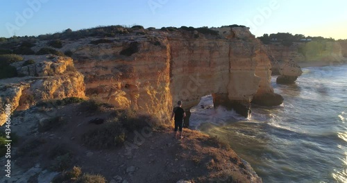 Praia Da Marinha A Natural Arch In Atlantic Coast With Person Standing On Clifftop In Lagoa, Portugal. - Aerial Overhead Shot