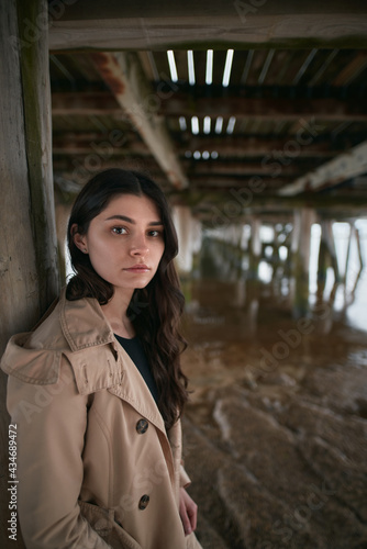 Portrait of stylish brunette under the wooden pier. Attractive and trendy girl with long brown hair by the seaside stand in tan color trench coat. © AlexGo