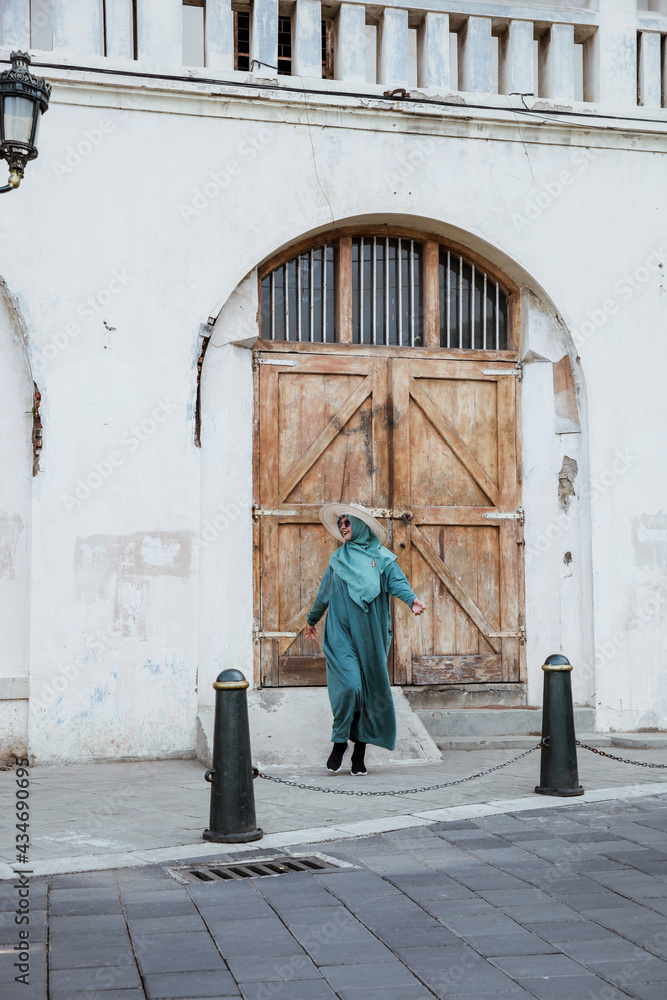 Happy Muslim Woman wearing green dress and summer hat walking on the street with old building on the background