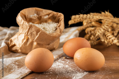 Close-up of eggs, bag of flour and ears of wheat, on wooden table and cloth, selective focus, black background, horizontal