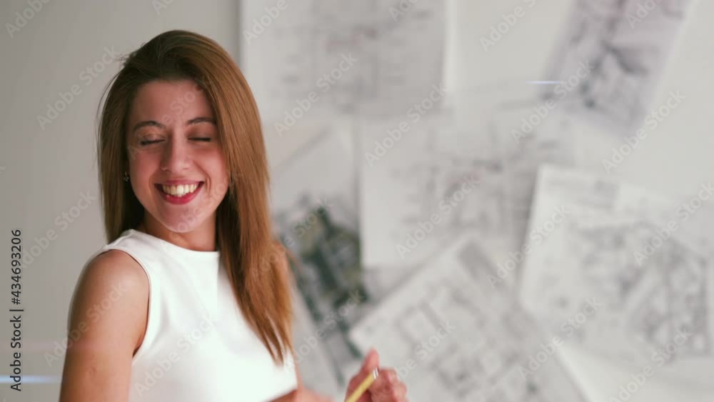 Beautiful portrait of young female designer standing in front of white wall of blueprints. Woman laughing, joking around while posing in front of her work. Concept of lifestyle, work. Beautiful portrait of young female designer standing in front of white wall of blueprints. Woman laughing, joking around while posing in front of her work. Concept of lifestyle, work.