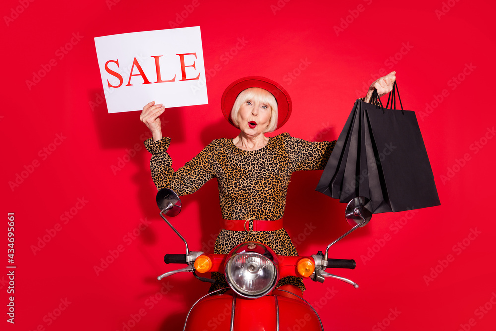 Photo of aged woman ride motorbike hold banner sale hold shopping bags black friday isolated over red color background Photo of aged woman ride motorbike hold banner sale hold shopping bags black friday isolated over red color background
