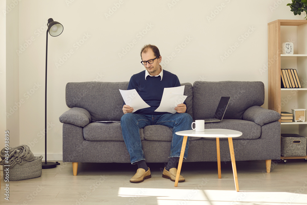 Senior citizen busy with paperwork at home. Mature man sitting on sofa with laptop computer and papers, looking through bills, reading pension plan documents or studying contract terms and conditions Senior citizen busy with paperwork at home. Mature man sitting on sofa with laptop computer and papers, looking through bills, reading pension plan documents or studying contract terms and conditions