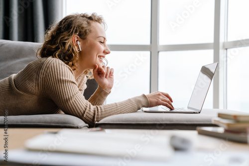 Young woman in earphones using laptop while lying on couch at home