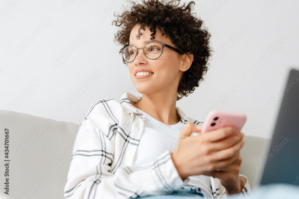 Young curly woman using cellphone and laptop while sitting on couch Young curly woman using cellphone and laptop while sitting on couch