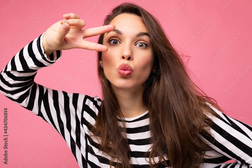 Obraz premium Photo of young european positive winsome sexy beautiful brunette woman with sincere emotions wearing casual striped pullover isolated on pink background with copy space and giving kiss