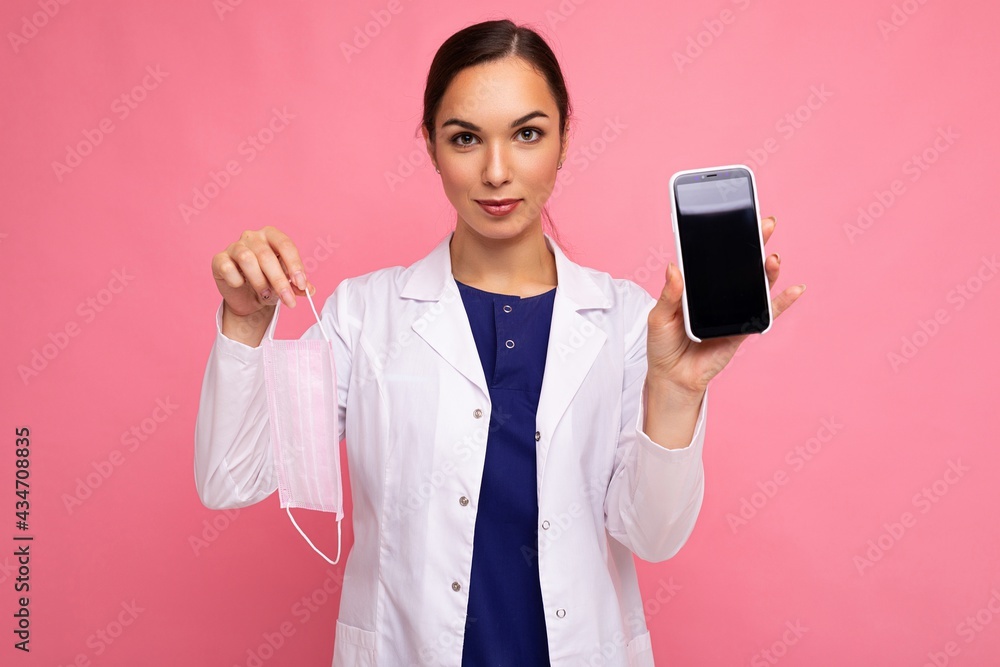 Attractive medical young woman wearing white coat holding mask showing modern mobile phone isolated over pink backgroung looking at camera Attractive medical young woman wearing white coat holding mask showing modern mobile phone isolated over pink backgroung looking at camera