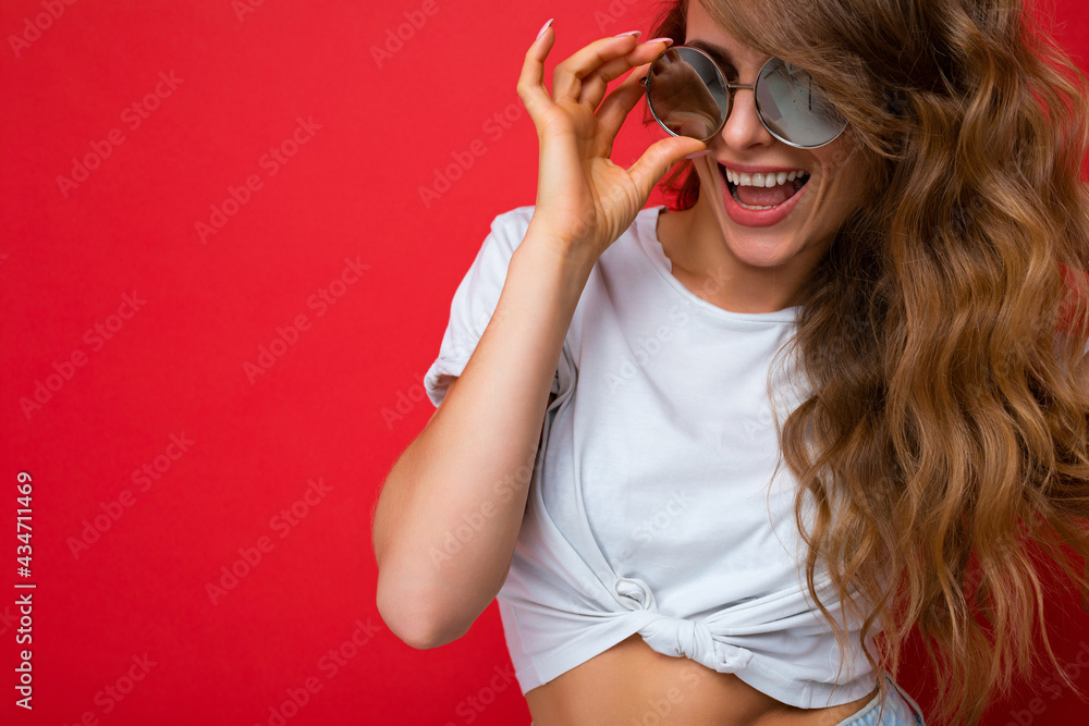 Closeup shot of beautiful smiling young dark blonde curly female person isolated over red background wall wearing casual white t-shirt and stylish sunglasses looking at camera Closeup shot of beautiful smiling young dark blonde curly female person isolated over red background wall wearing casual white t-shirt and stylish sunglasses looking at camera