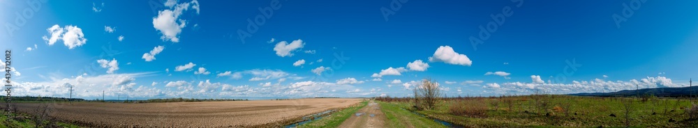 Panorama of plowed field in spring, blue sky, green grass, field road.