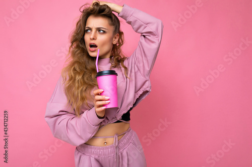 Photo shot of beautiful young shocked blonde woman wearing pink sport clothes isolated over pink background holding paper coffee cup for mockup drinking and looking to the side
