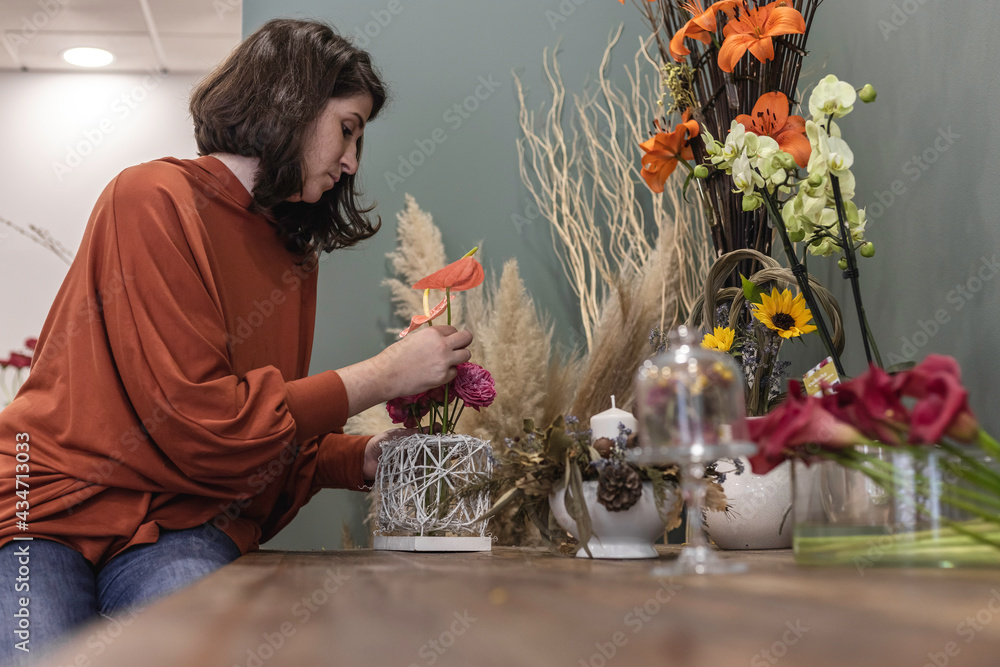 View of a young woman working as florist in her flower shop, sitting at wooden table and creating flower decoration, businesswoman concept. - obrazy, fototapety, plakaty 