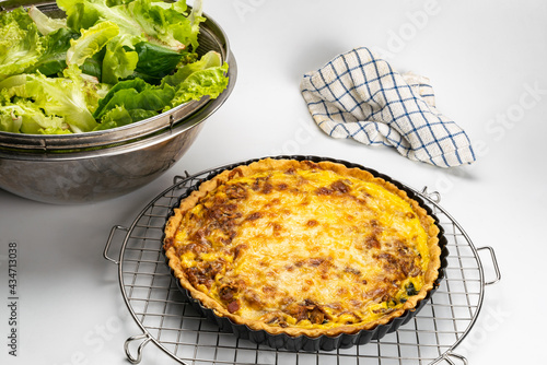 Freshly baked quiche in black metal oven pie baking plate on a metal cooling rack with various vegetable and tablecloth on white background.