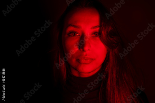 Young pretty woman looks at camera in contrast red light. Sensual female person posing in dark studio with red neon lightning. Portrait of calm woman, abuse addiction, depression, imprisonment concept