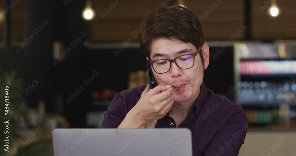 Asian man with laptop having a snack and talking on smartphone while sitting at a cafe Asian man with laptop having a snack and talking on smartphone while sitting at a cafe