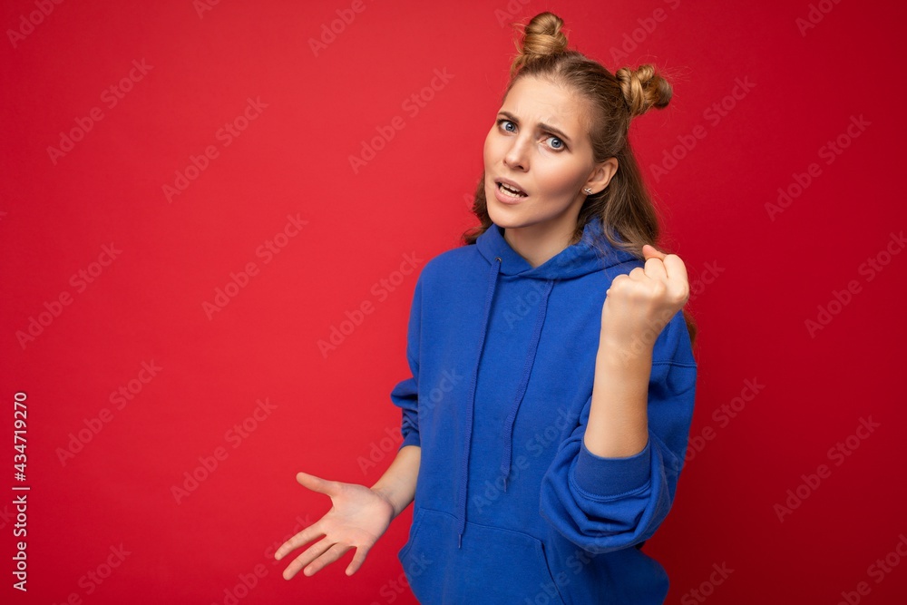 Photo of emotional agressive young beautiful blonde woman with two horns with sincere emotions wearing hipster bright blue hoodie isolated over red background with free space and showing fist Photo of emotional agressive young beautiful blonde woman with two horns with sincere emotions wearing hipster bright blue hoodie isolated over red background with free space and showing fist