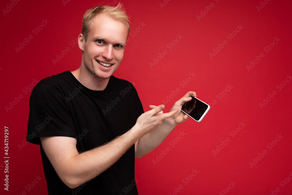 Shot of handsome smiling blonde young male person wearing black t-shirt standing isolated on red background with copy space holding smartphone showing phone in hand with empty display for mockup Shot of handsome smiling blonde young male person wearing black t-shirt standing isolated on red background with copy space holding smartphone showing phone in hand with empty display for mockup