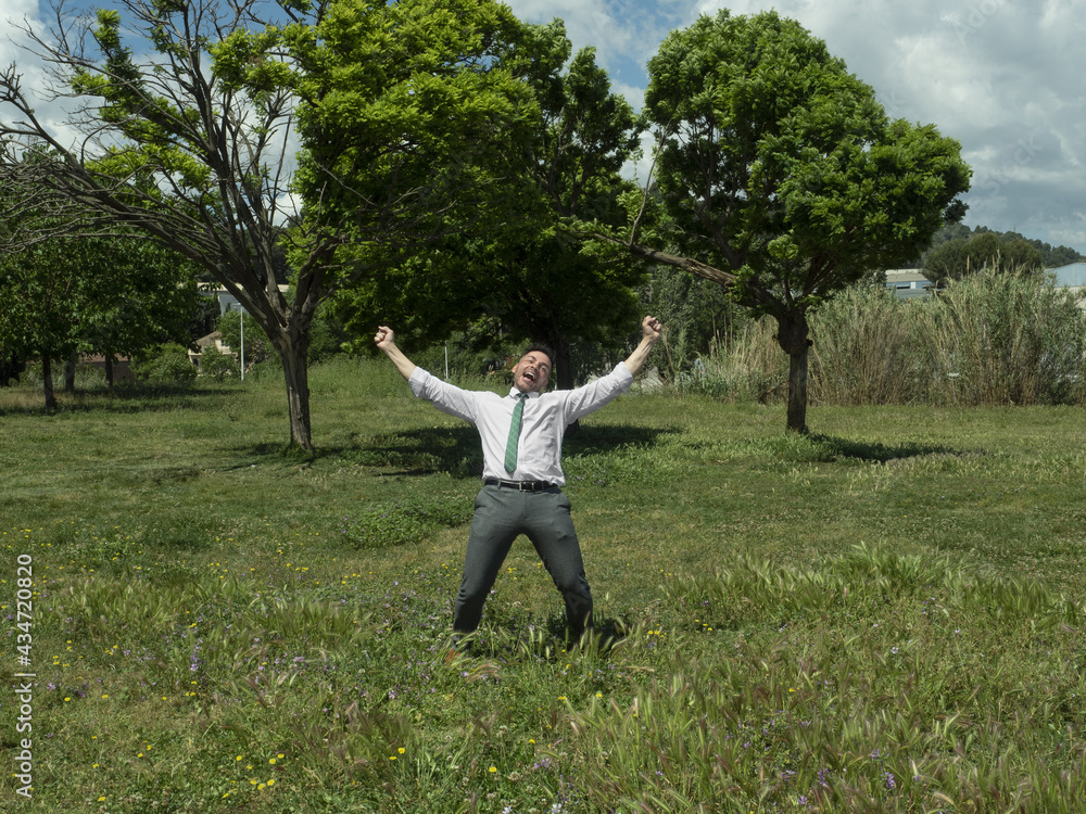 a green park with trees a businessman with a positive attitude is celebrating with open arms and a very happy face.