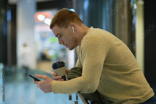 young man using smartphone and listens to music in bluetooth headphones. lonely man in autumn jacket drinks coffee.