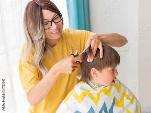 Mother cuts her son's hair. Little boy sits, covered with cloth, and holds pair of scissors. New normal in quarantine of coronavirus COVID-19. Self care at home lifestyle.