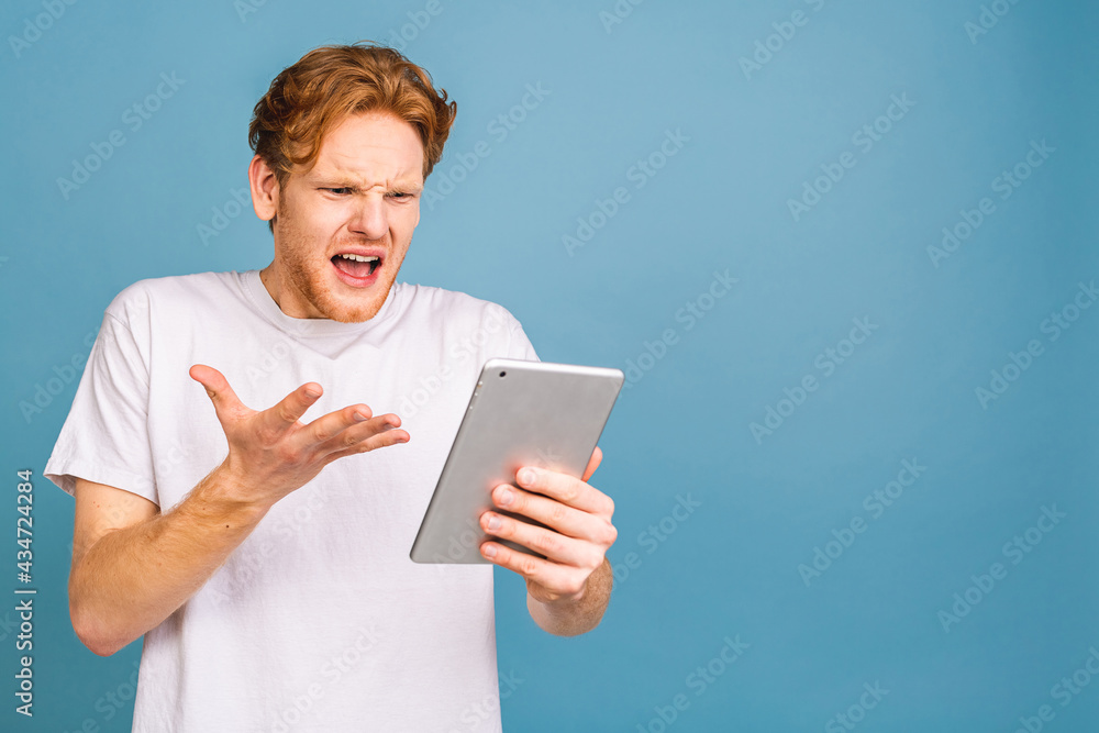 Happy shocked amazed young bearded red-haired man in casual using tablet and standing isolated over blue background. Happy shocked amazed young bearded red-haired man in casual using tablet and standing isolated over blue background.