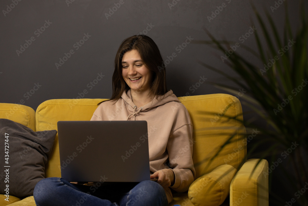 Cheerful laughing Attractive long-haired brunette girl Chatting and texting on laptop sitting on yellow sofa in front of potted dracena. Communicating online, using Internet chat, messenger Cheerful laughing Attractive long-haired brunette girl Chatting and texting on laptop sitting on yellow sofa in front of potted dracena. Communicating online, using Internet chat, messenger