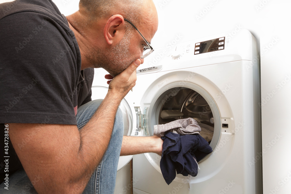 Obraz premium bald man in gray jeans and t-shirt sits in front of a washing machine at home. Load the washing machine with dirty clothes. Bright and spacious living room with a modern interior.