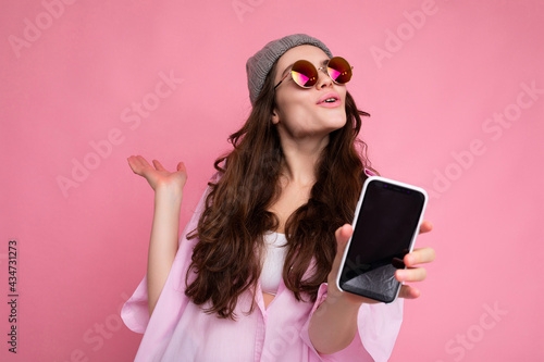 Positive attractive young brunette female person wearing stylish pink shirt grey hat and colourful sunglasses isolated over pink background holding in hand and showing mobile phone with empty display