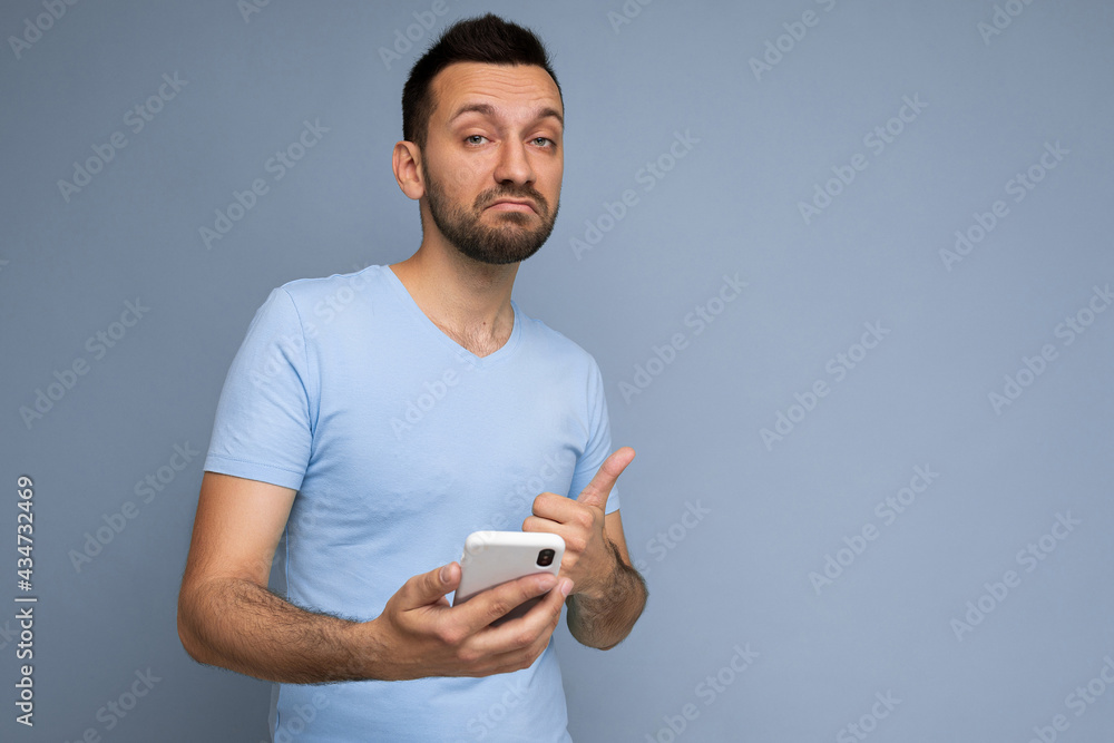 Thoughtful handsome young man isolated over background wall wearing everyday clothes holding and using mobile phone writing sms looking at camera and thinking Thoughtful handsome young man isolated over background wall wearing everyday clothes holding and using mobile phone writing sms looking at camera and thinking