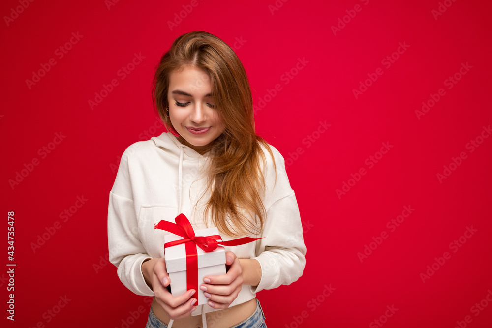Photo shot of pretty positive surprised young dark blond woman isolated over colourful background wall wearing trendy outfit look holding gift box and looking at present box with red ribbon Photo shot of pretty positive surprised young dark blond woman isolated over colourful background wall wearing trendy outfit look holding gift box and looking at present box with red ribbon