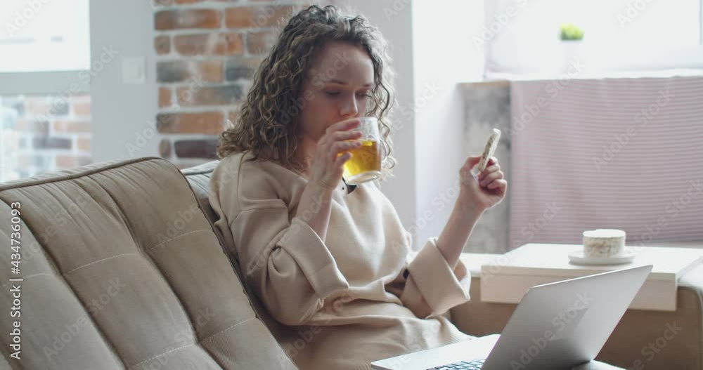 Beautiful cheerful girl using silver laptop while sitting on sofa against window in living room at home.Relax and laughing. High quality 4k footage