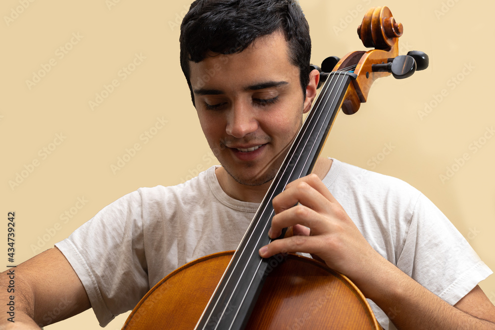 Young teenage girl smiling and playing fretted string musical instrument Cello placing notes with left hand wearing short white T-shirt with light yellow background. Young teenage girl smiling and playing fretted string musical instrument Cello placing notes with left hand wearing short white T-shirt with light yellow background.
