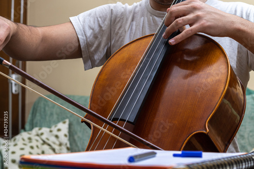 Detail of a teenager playing the classical music instrument Chelo during an online class with a notebook and pen next to him to take notes with selective focus