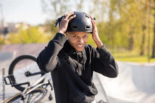 A young boy sits on a ramp at a skatepark with his bike lying wheel up behind him. The guy has finished riding and takes his black helmet off his head and slides it off with two hands