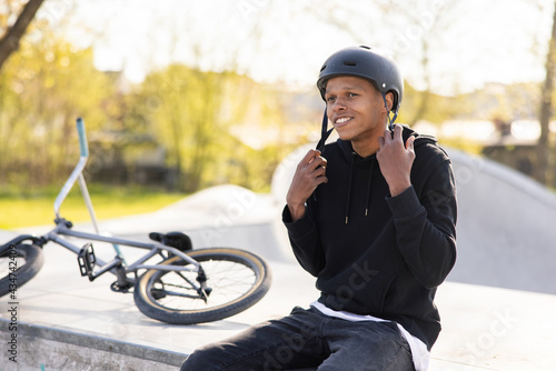 A boy who has fallen off bike, a bmx, and is sitting on a concrete ramp, gets ready to ride back on the track with friends, puts helmet on his head and fastens it under neck
